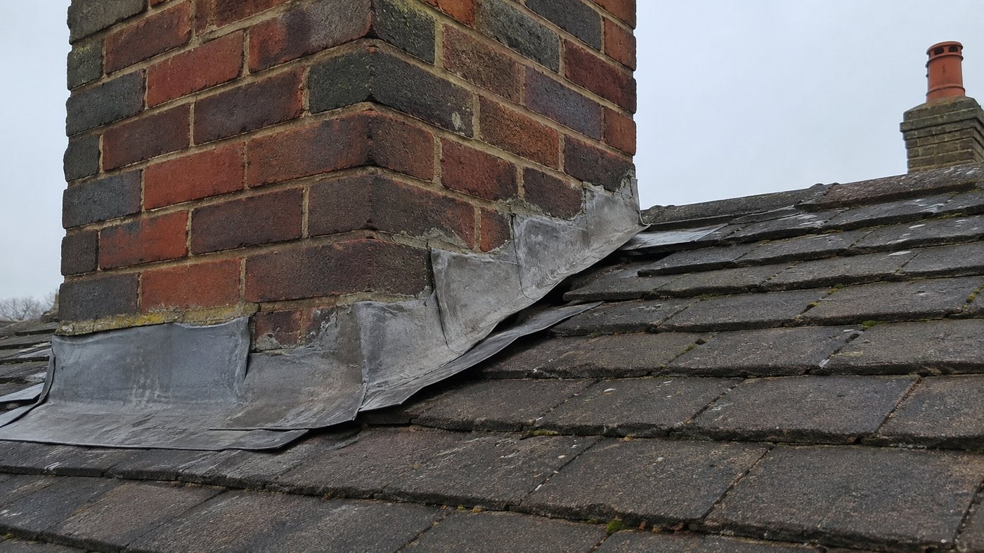 Worn lead flashing around the base of a brick chimney on a tiled roof.